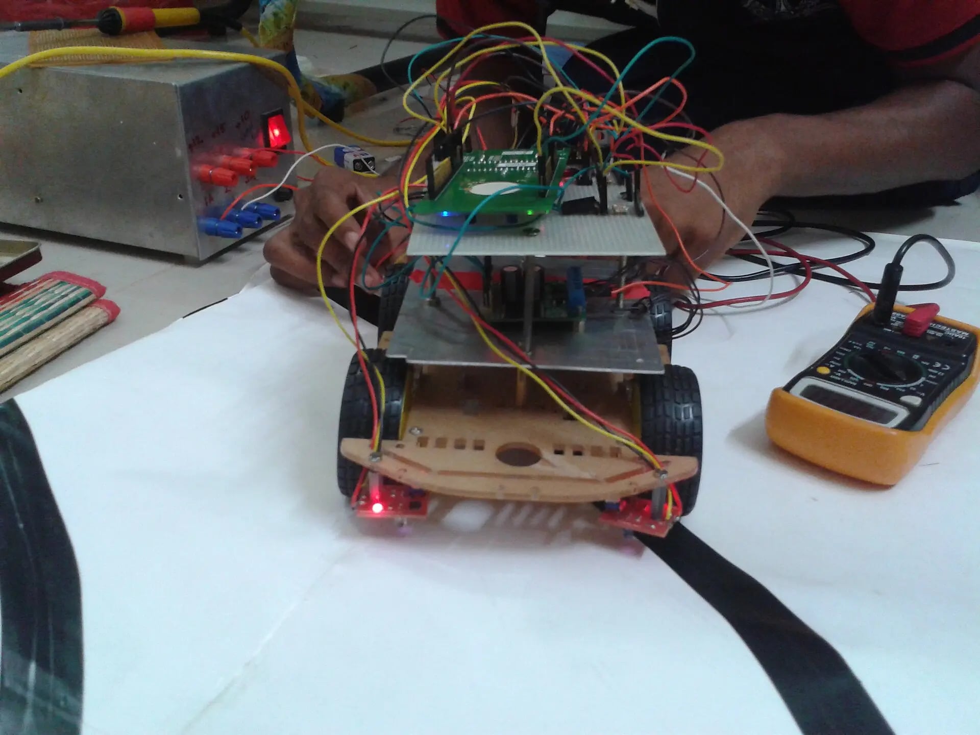 A two-deck robot car under test on a lab bench, wired to a bench power supply with a multimeter beside it.
