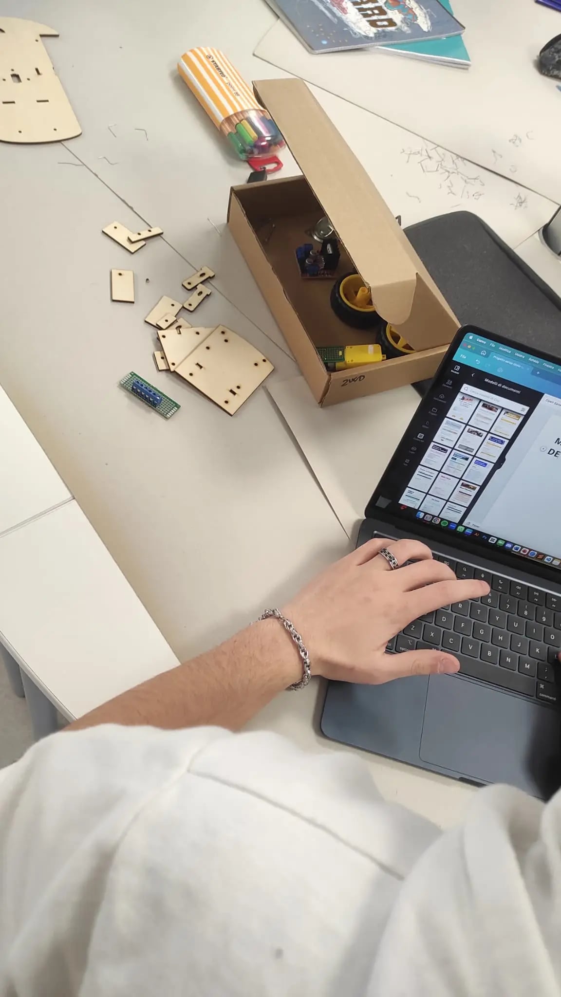 Overhead view of a bench: laser-cut plywood chassis parts, a kit box of motors and wheels, and a student working at a laptop.